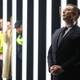 An image of Elon Musk in a black suit and dark tie, standing with his hands clasped in front and head raised, eyes closed. In the background are consecutive vertical white and black bars, three blurry figures stand in between them.